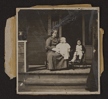 A monochromatic snapshot with surface damage and remnants of adhesive pressure senstive tape shows an older woman seated on the front porch of a home. An infant is seated on her left knee and another older child is seated on a tricycle to the woman's left.