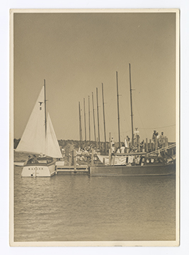 The Fort Worth Boat Club sailboat, The Ranger, is viewed from behind at a dock with its white sails raised.. Several men and the masts of other sailboats are visible at the dock to the right.