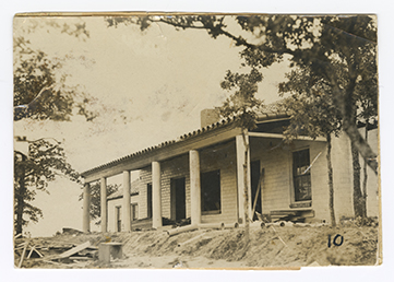 A wide unfinished building, with a covered porch supported by six columns is viewed left of center. Bare dirt and loose construction materials are visible to the front and sides of the building.