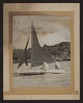 A small sailboat in full sail on the water is viewed from the right, or starboard, side. One man, seated on the middle right side of the hull, has his back to the camera but is looking towards the camera over his left shoulder. A second man is seated at the back right of the hull, also with his back towards the camera, and is looking in the direction the boat is sailing. He is steering the boat with the rudder. The photograph is mounted to a tan board with old tape and has stains on the upper half of the image. A horizontal crack is running through the surface of the image across the two men's faces.